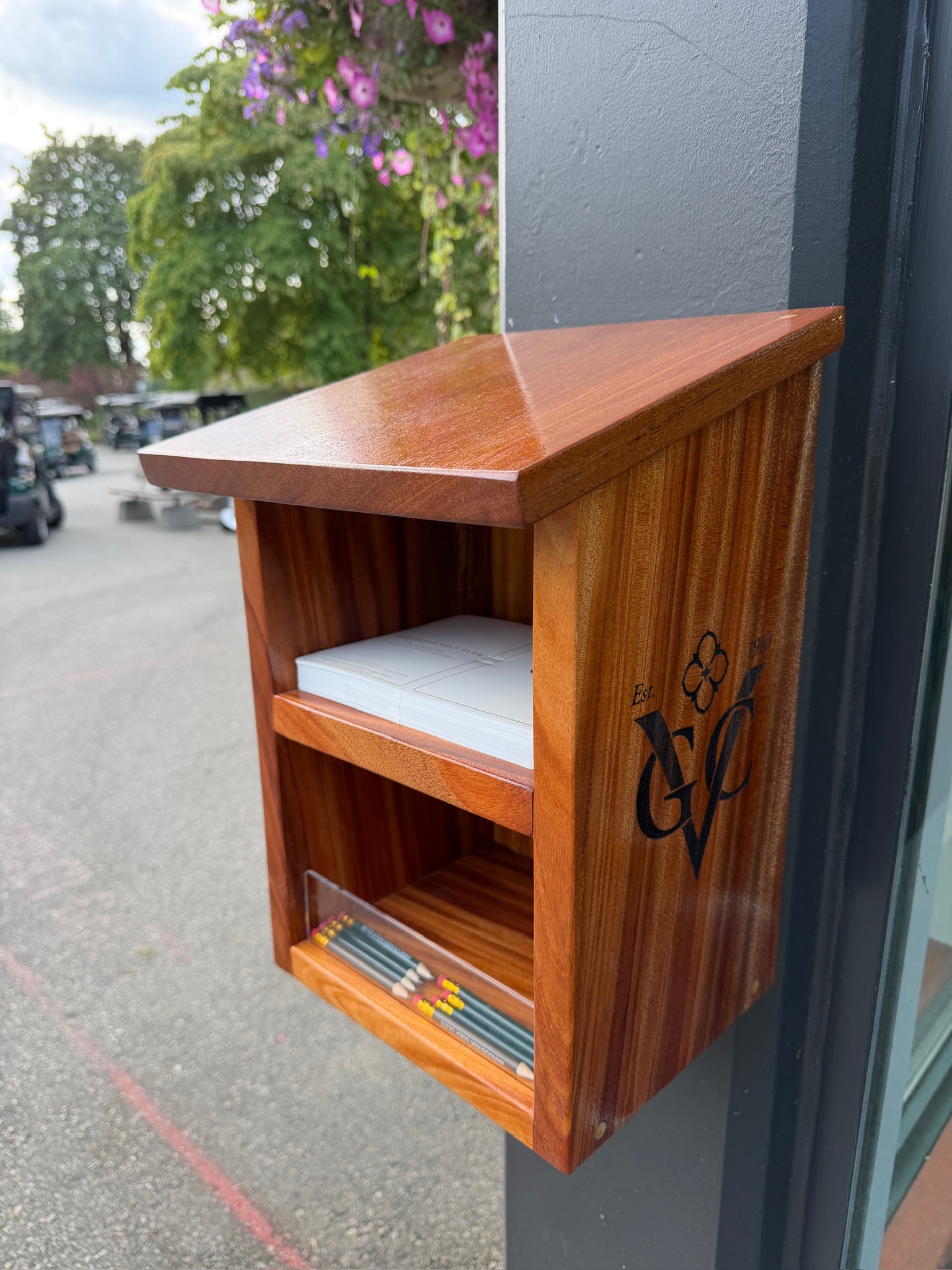 Wall-mounted wooden scorecard and pencil station with shelved compartments and engraved club crest, installed at a pro shop entrance.