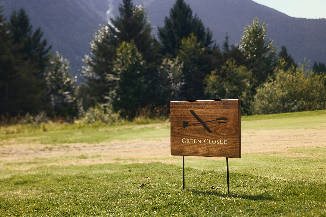 Wooden Green Closed course sign with engraved crossed putter and cup graphic, mounted on steel stakes with a mountain and forest backdrop.