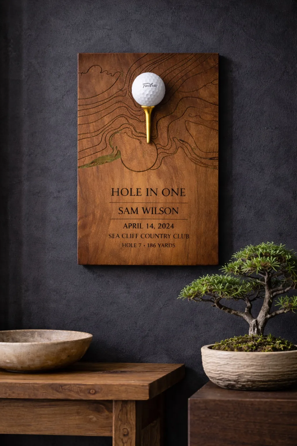 Walnut hole-in-one plaque with engraved topographic contour lines, golf ball on gold tee, mounted on a dark wall.