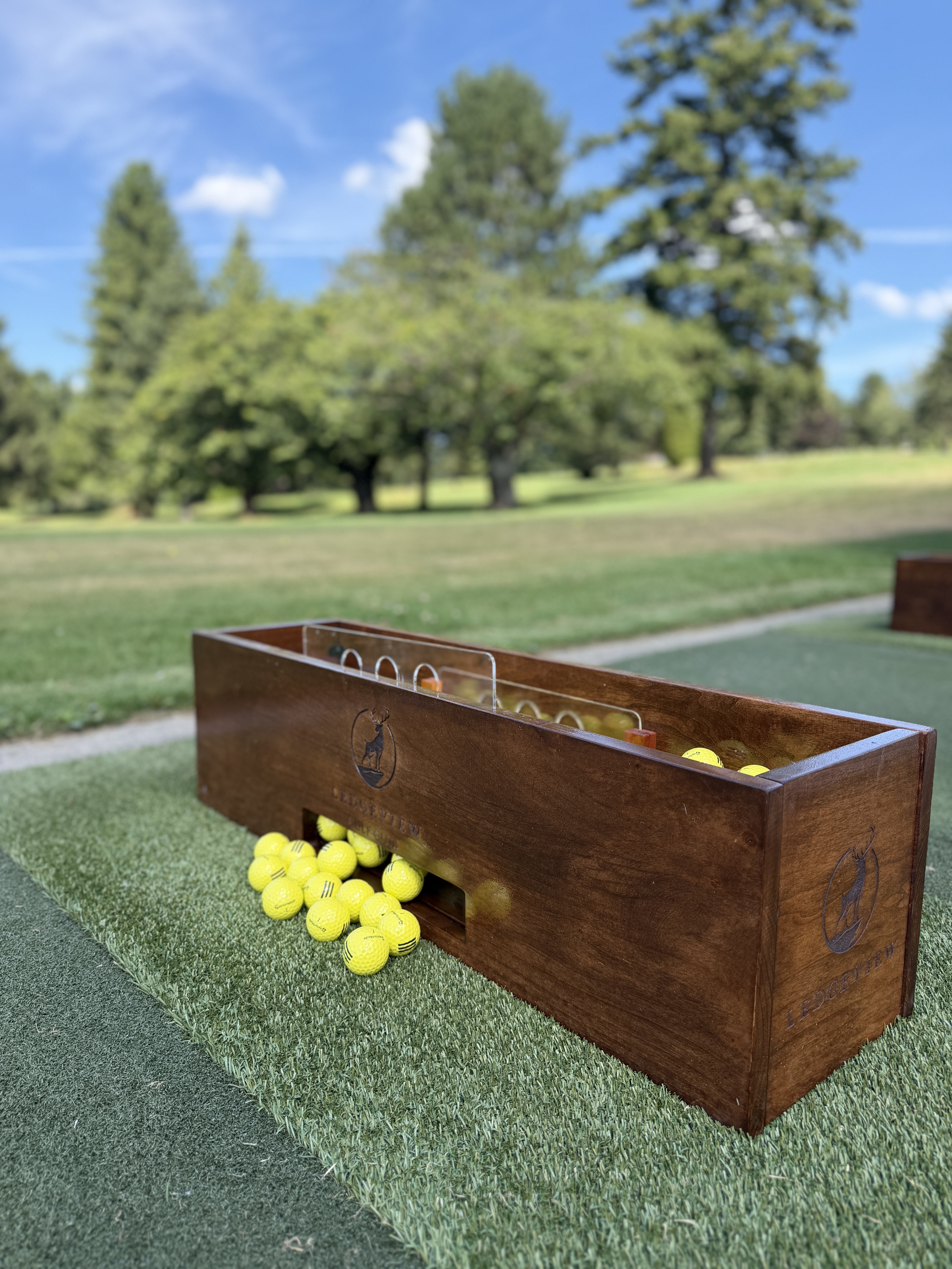 Walnut driving range ball dispenser on a range mat with yellow range balls, featuring engraved Ledgeview Golf Club logo.