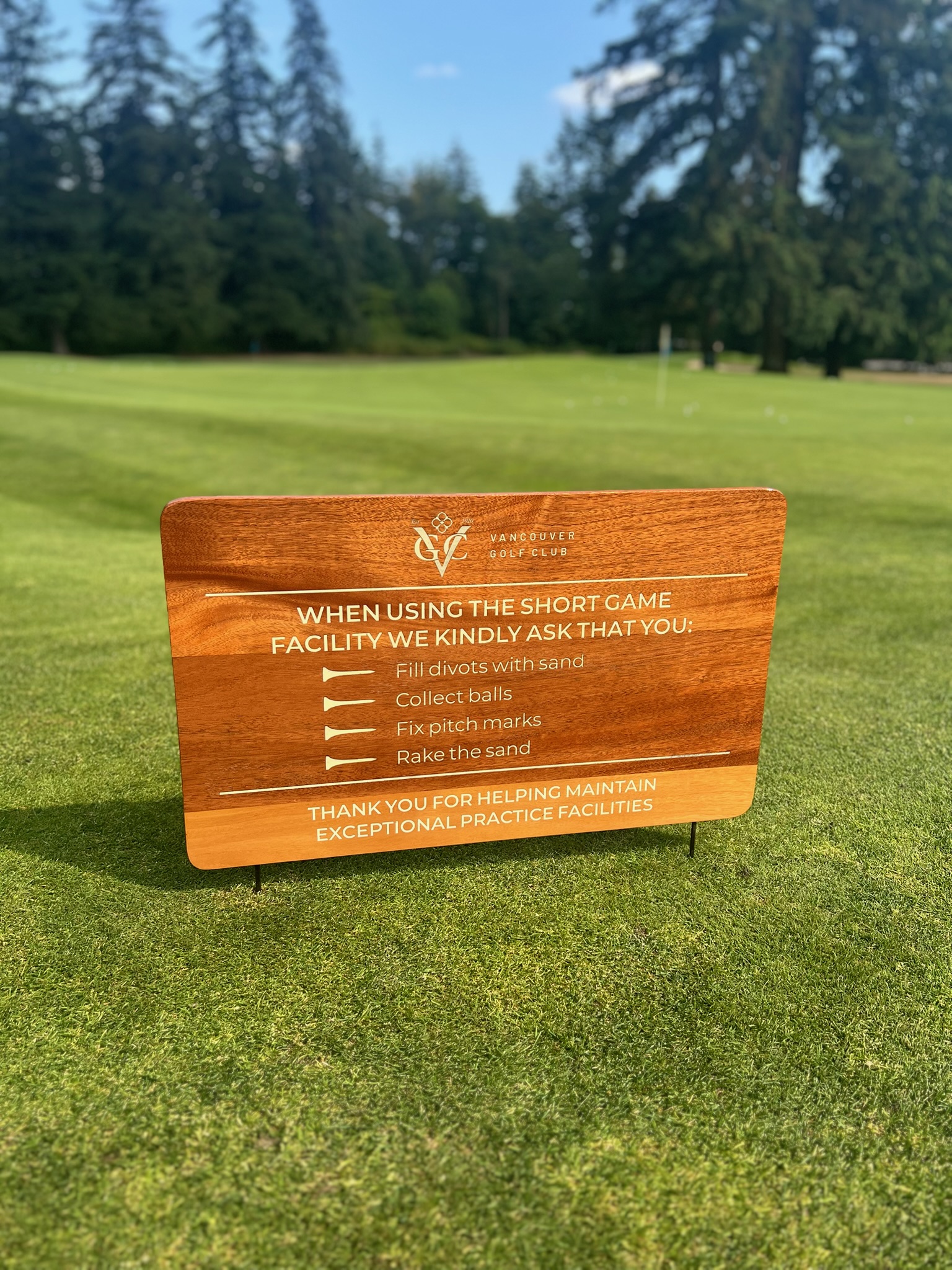 Walnut short game facility etiquette sign with Vancouver Golf Club logo and engraved guidelines for divot repair, ball collection, and sand raking, mounted on steel stakes on a practice green.