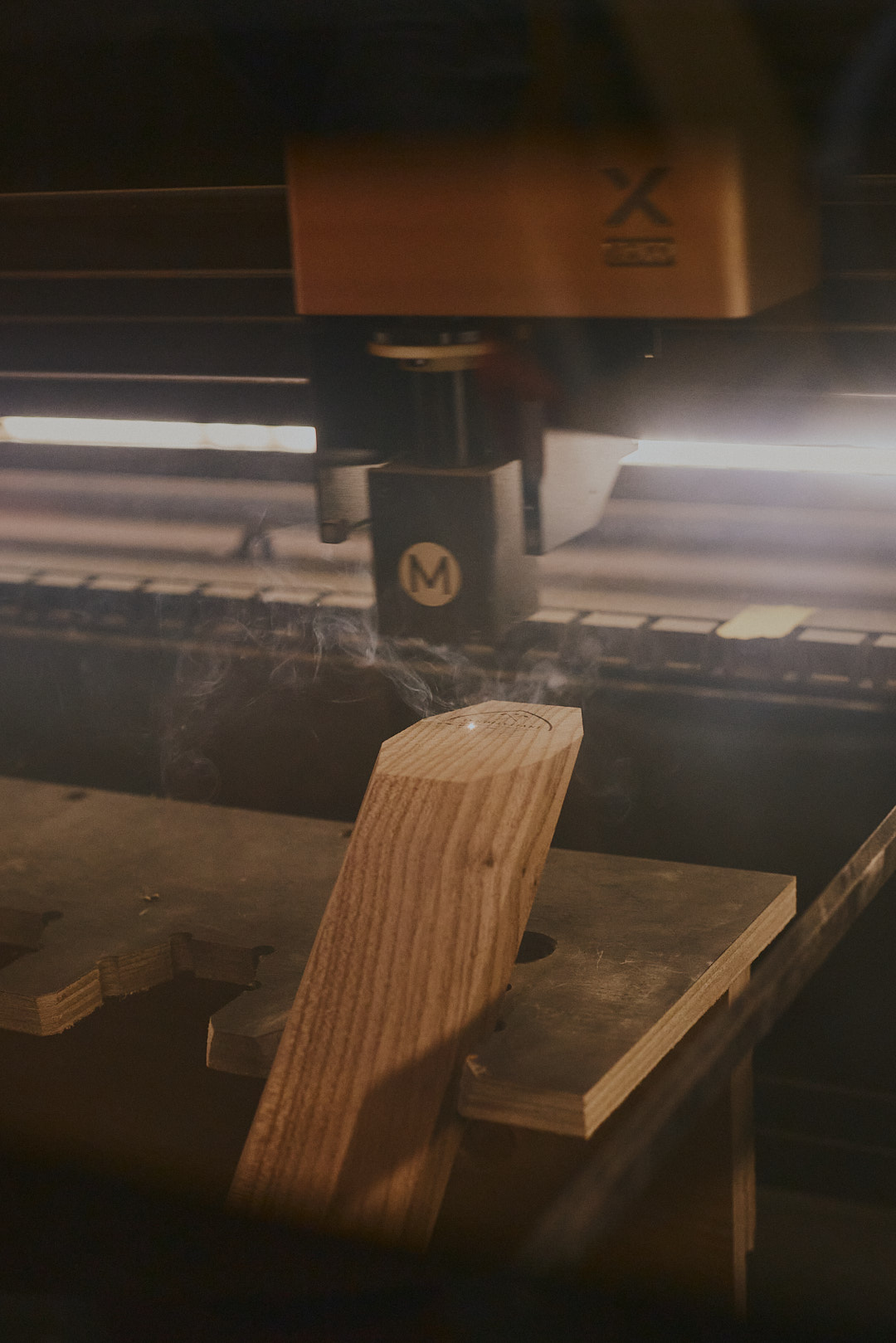 Laser engraving a tee marker in the studio, smoke rising as the engraving head works across the wood surface.